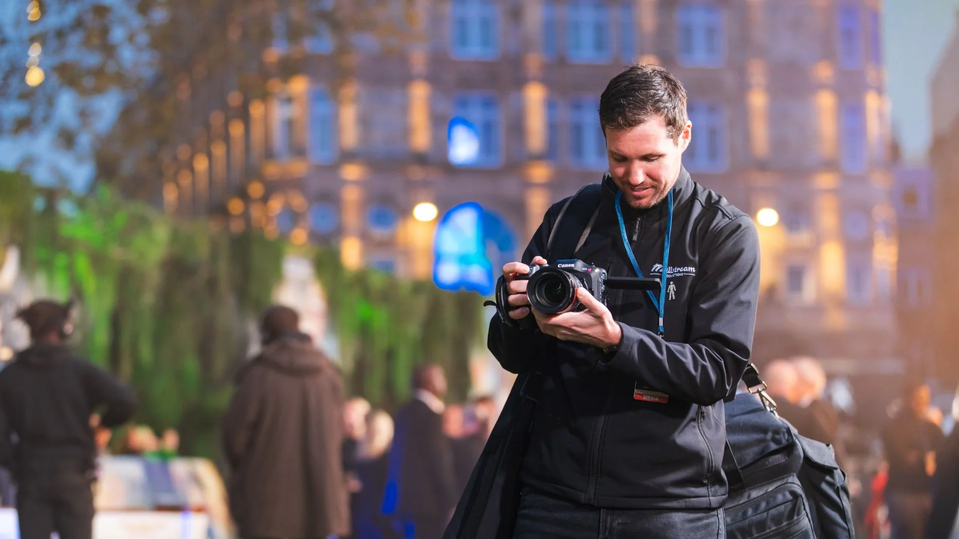 Photographer in dark jacket reviewing camera screen at a busy outdoor event.