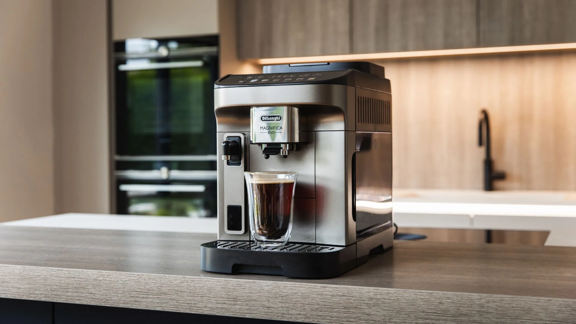 Silver coffee machine dispensing dark coffee into a glass in a modern kitchen.