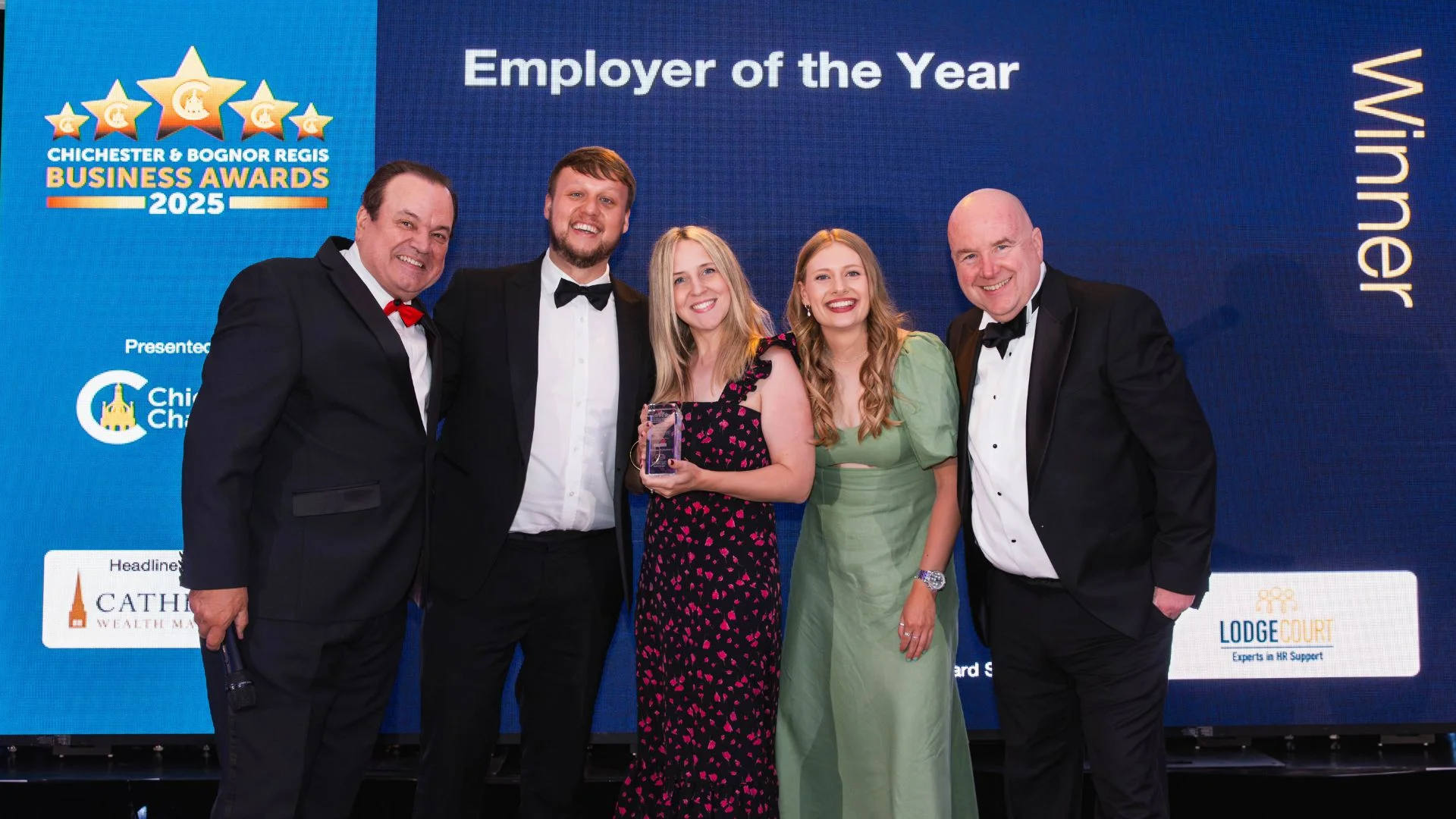 Five smiling people, three men and two women, holding a business award trophy on stage.
