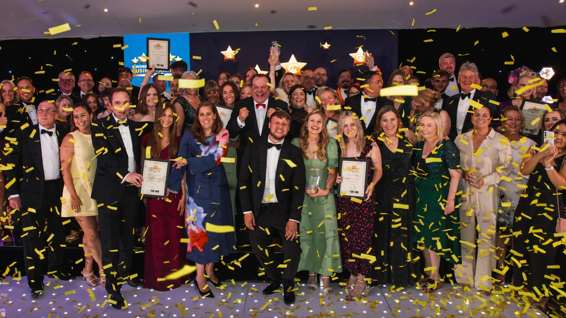 Large group in formal wear celebrating, surrounded by falling gold confetti at an awards ceremony.