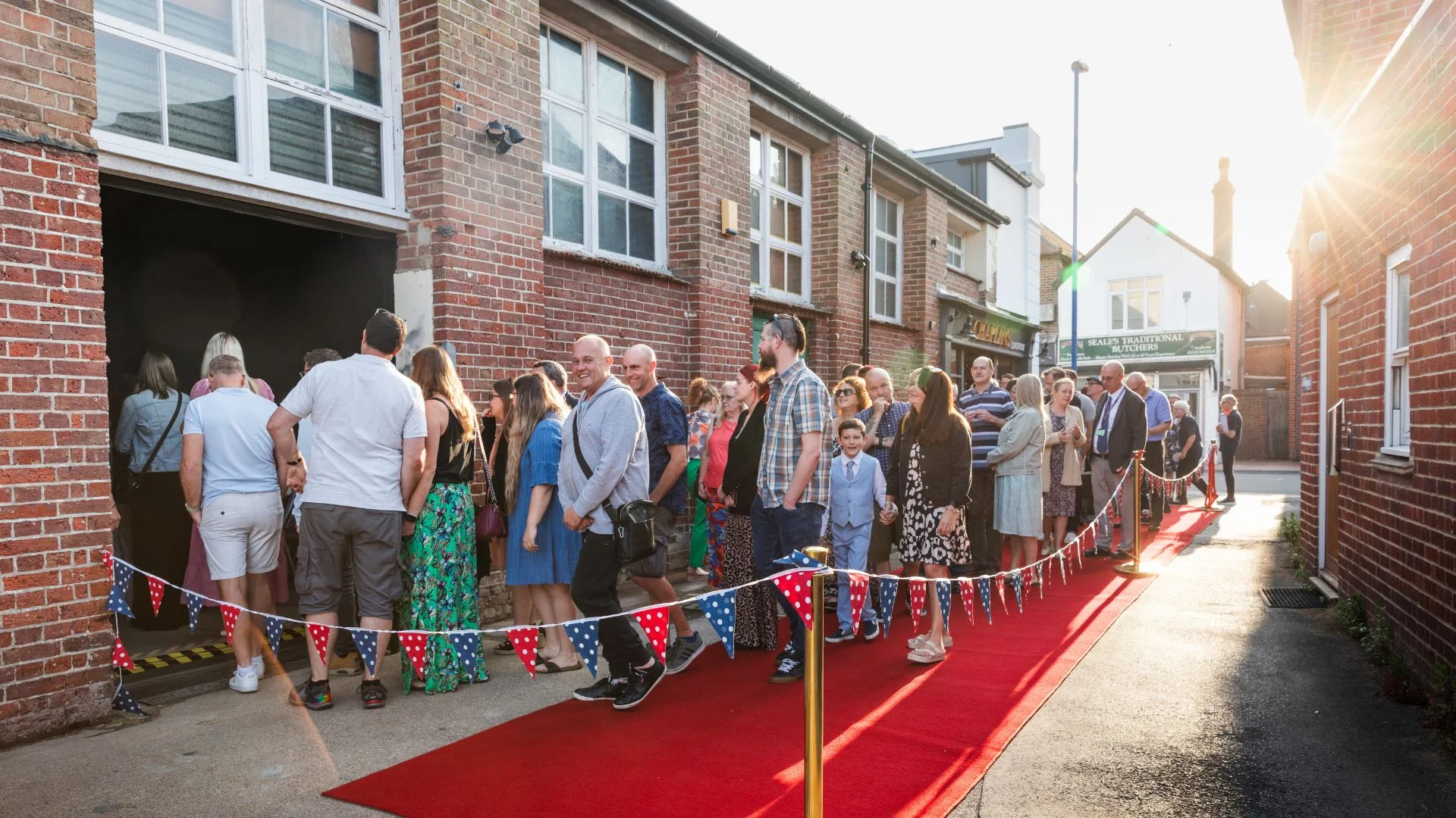 People queue on a red carpet with Union Jack flags outside a brick building.