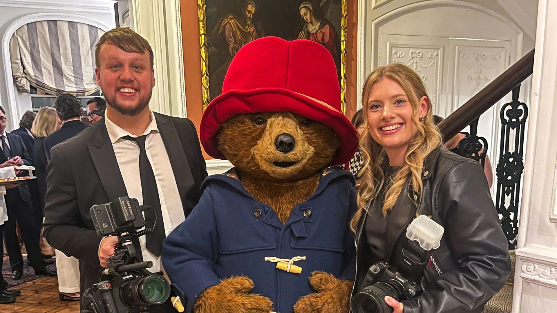 Man, Paddington Bear, and woman holding cameras smile at an event.