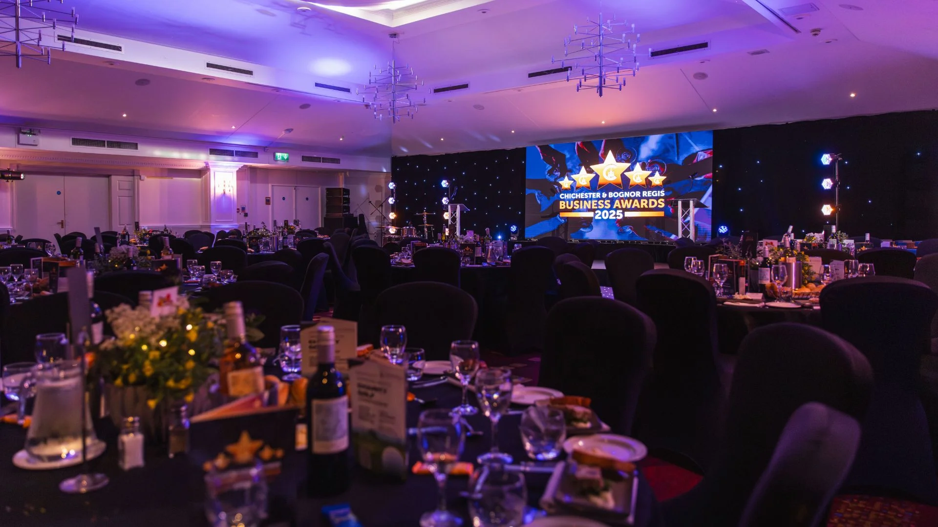 Gala dinner event in a ballroom with tables, chairs, and a large awards screen in purple light.