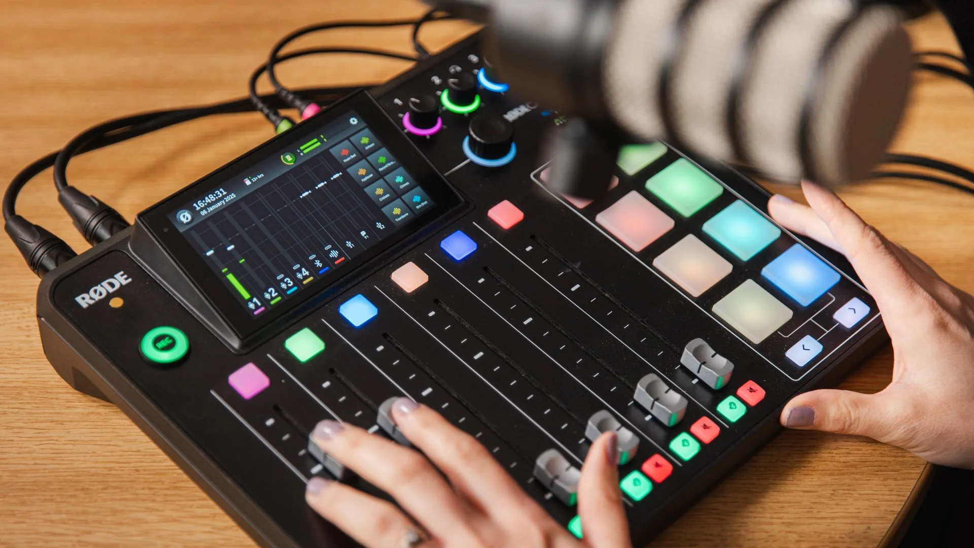 Hands operate a RODE podcast mixer with faders, screen, and colourful pads.