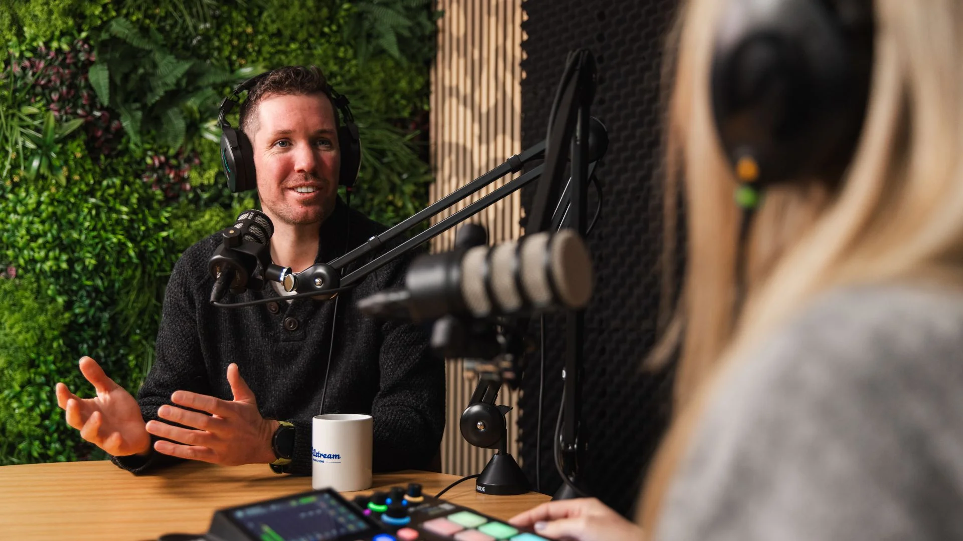 Smiling man in headphones gestures while recording a podcast with a blonde engineer.