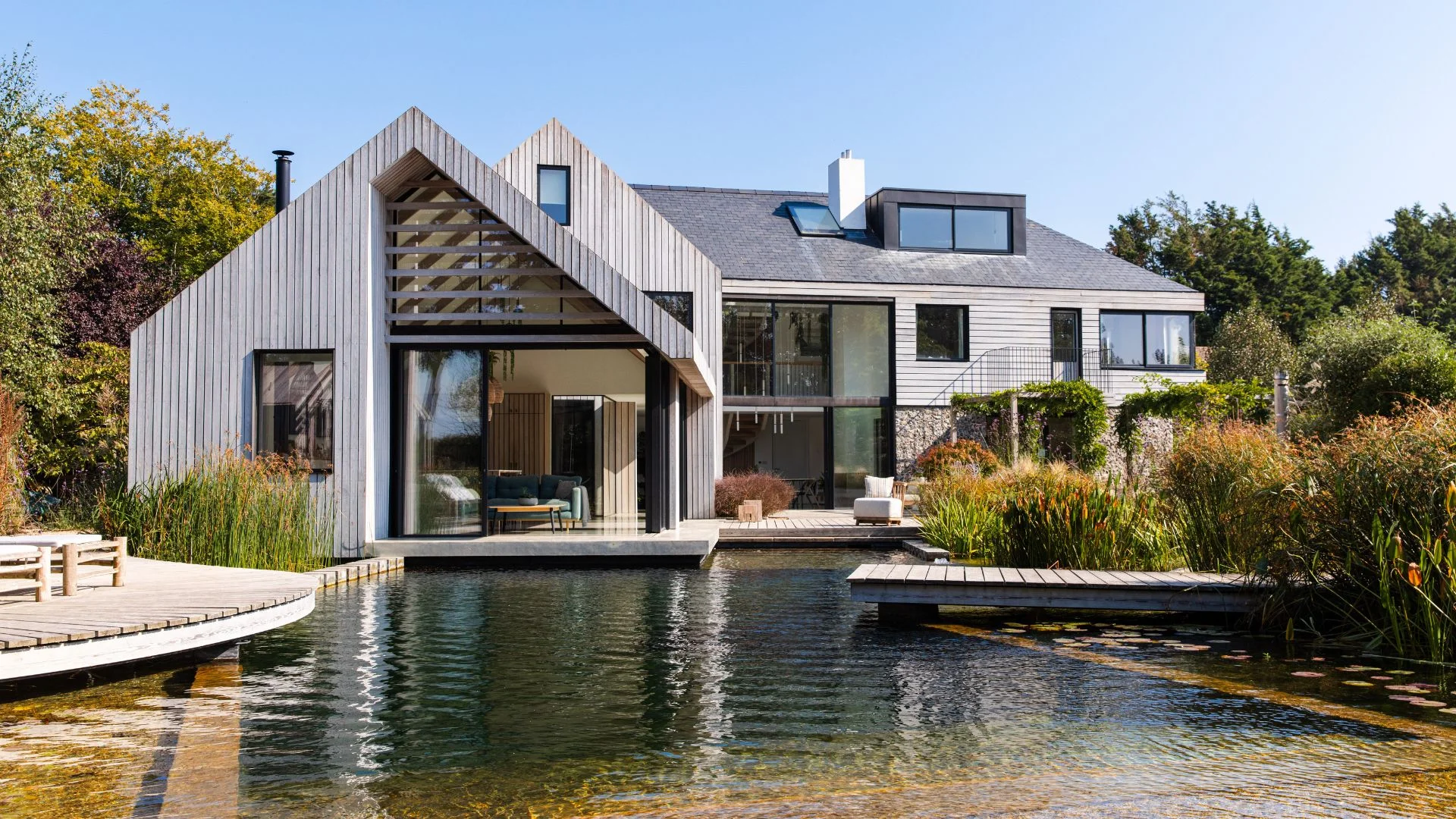 Modern house with light wood cladding and dark roof overlooking a large reflective pond.