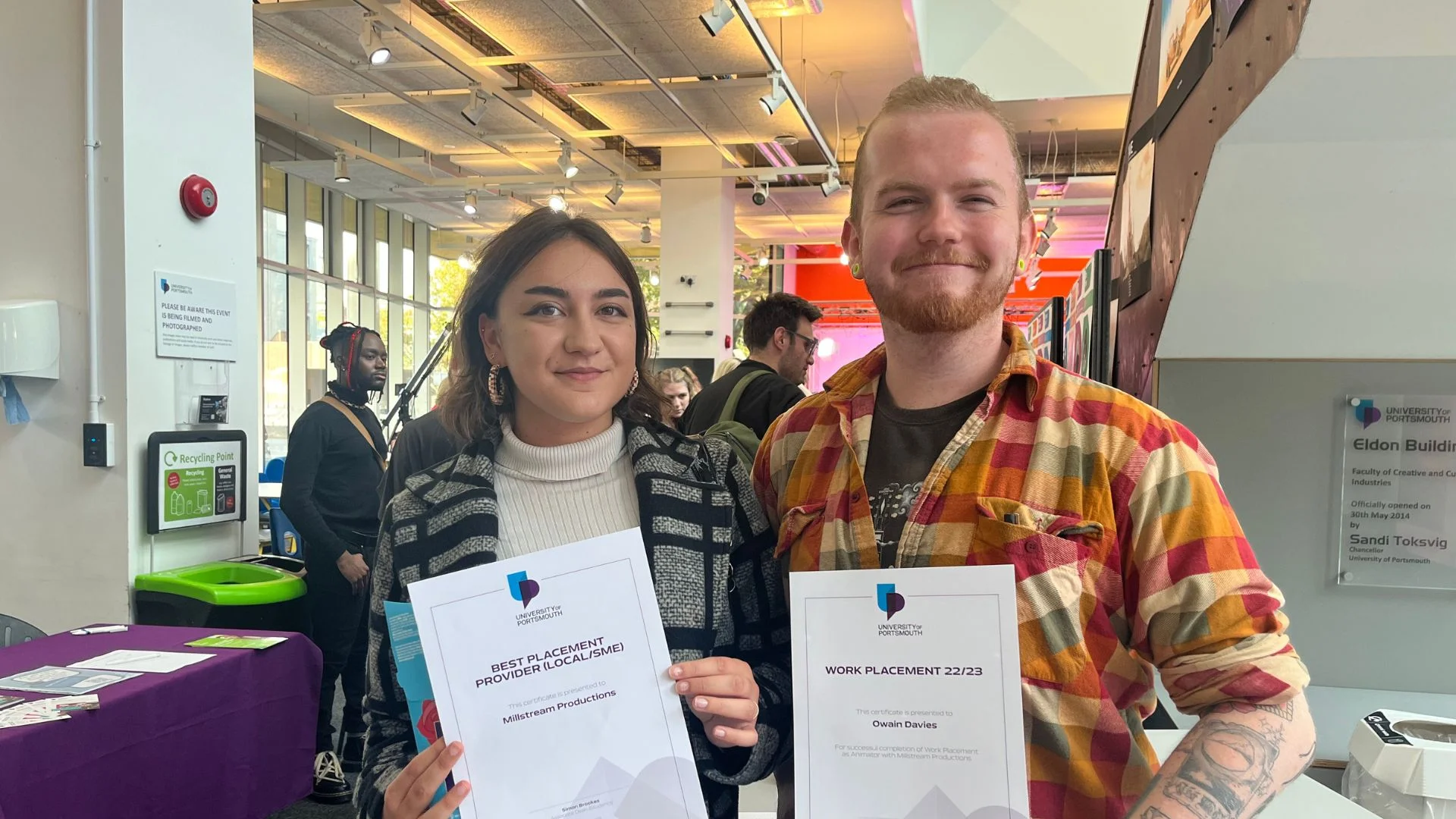 Photograph: Two smiling young people proudly hold certificates at an indoor event.