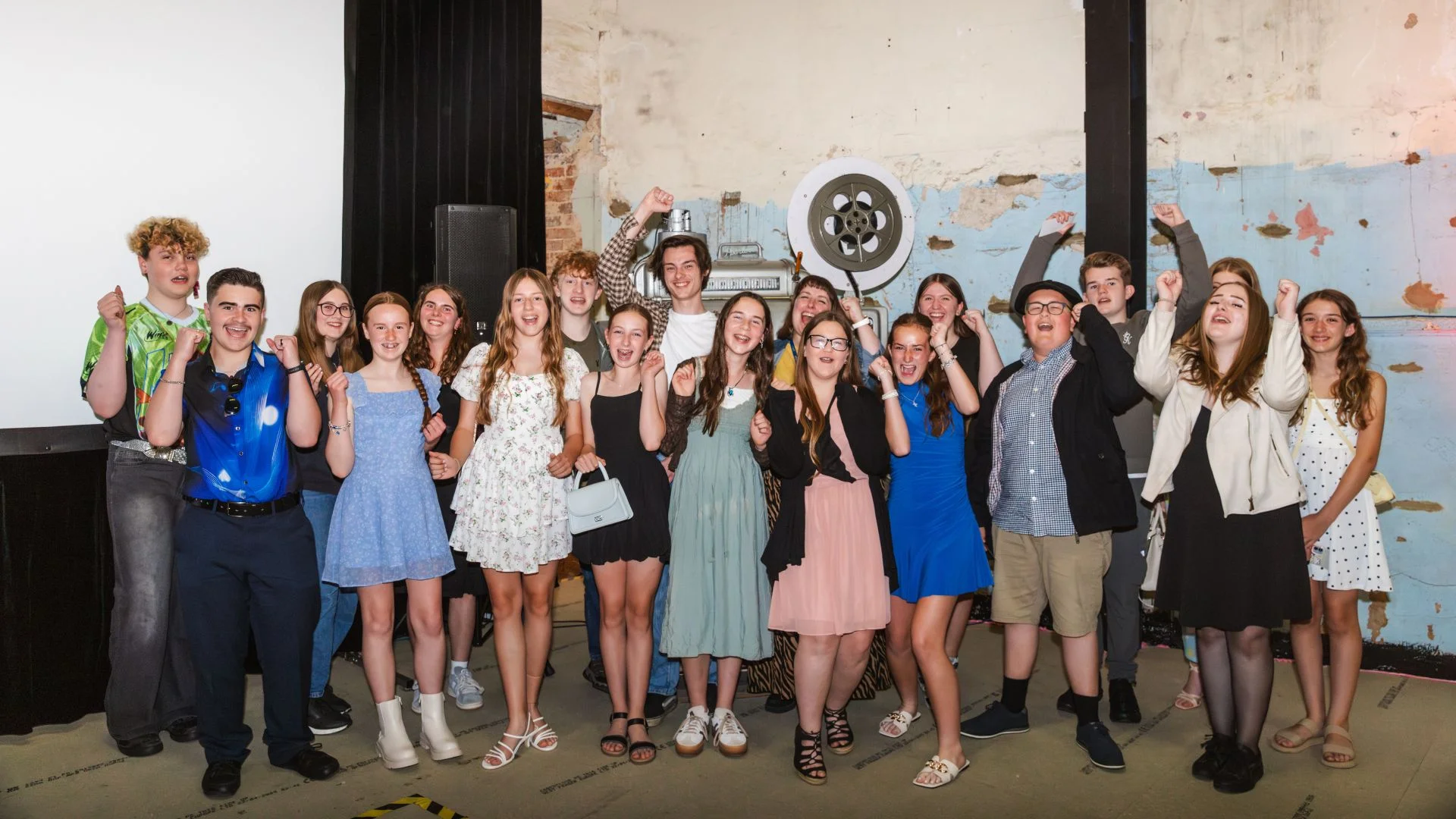 Photograph: Group of smiling teenagers celebrate, fists raised, in a casual event hall.
