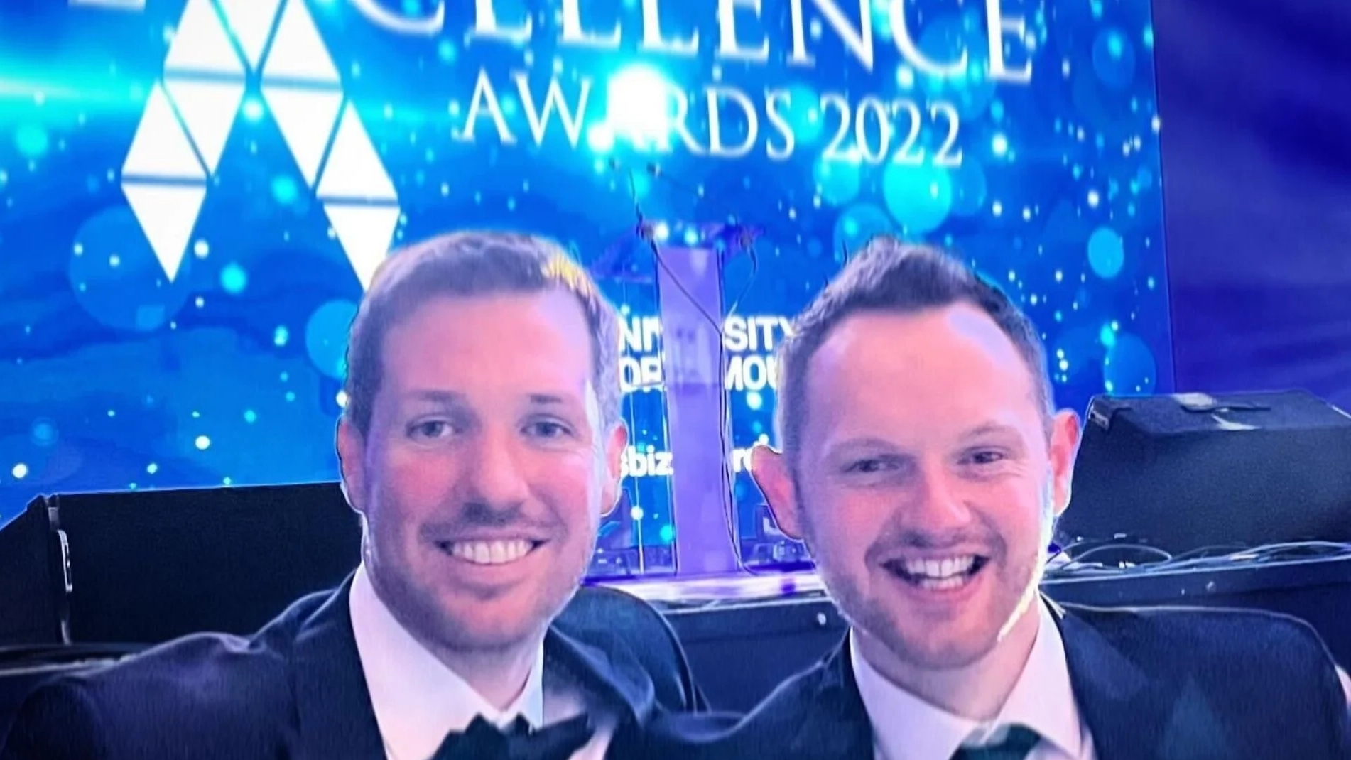 Two smiling men in suits at the Excellence Awards 2022.