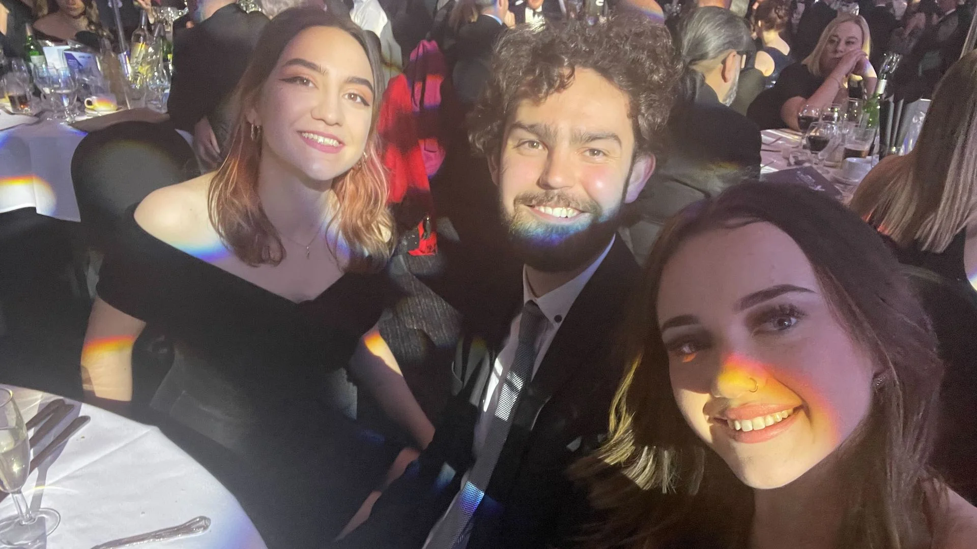 Three smiling people at a formal dinner, with rainbow light reflections on their faces.