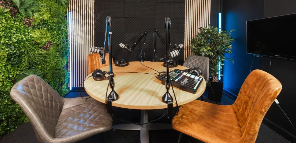 Photograph: Modern podcast studio with round table, two chairs, mics, mixing desk, and plant wall.