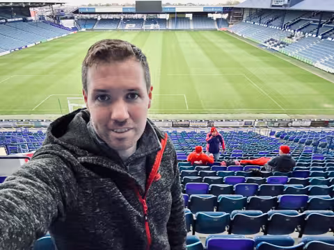 Man in a grey hoodie taking a selfie in a large, mostly empty football stadium.