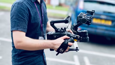 Man holding a professional video camera rig, blurred blue car in background.