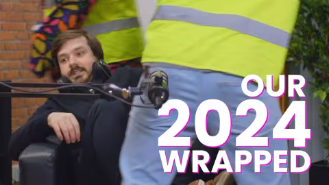 Man in black looks amused as person in high-vis walks past with camera rig.