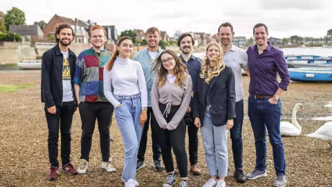 Photograph: Eight smiling people stand on a riverbank with boats and swans nearby.