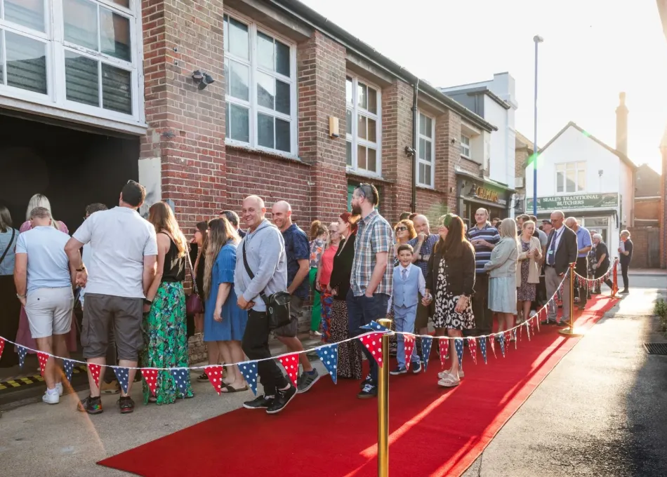 Photograph: Attendees queue on a red carpet with bunting leading to a building. Golden hour.