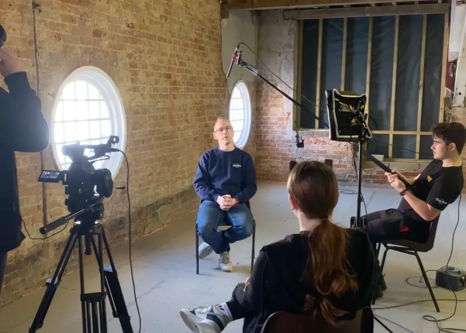 Film crew recording an interview in a brick-walled room with round windows.