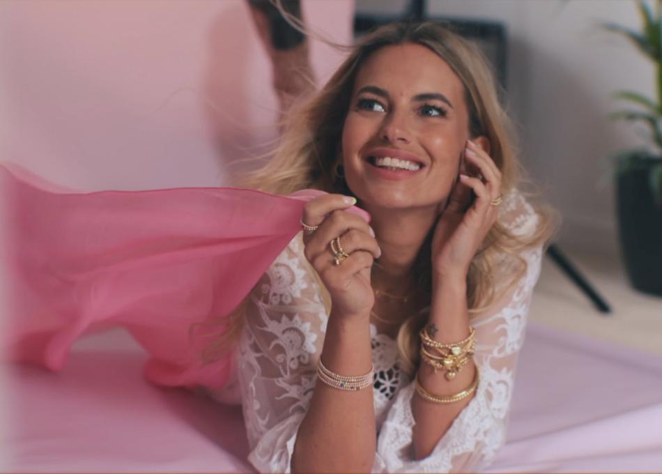 Smiling blonde woman in a lace top, lying on a pink studio background with gold jewellery.