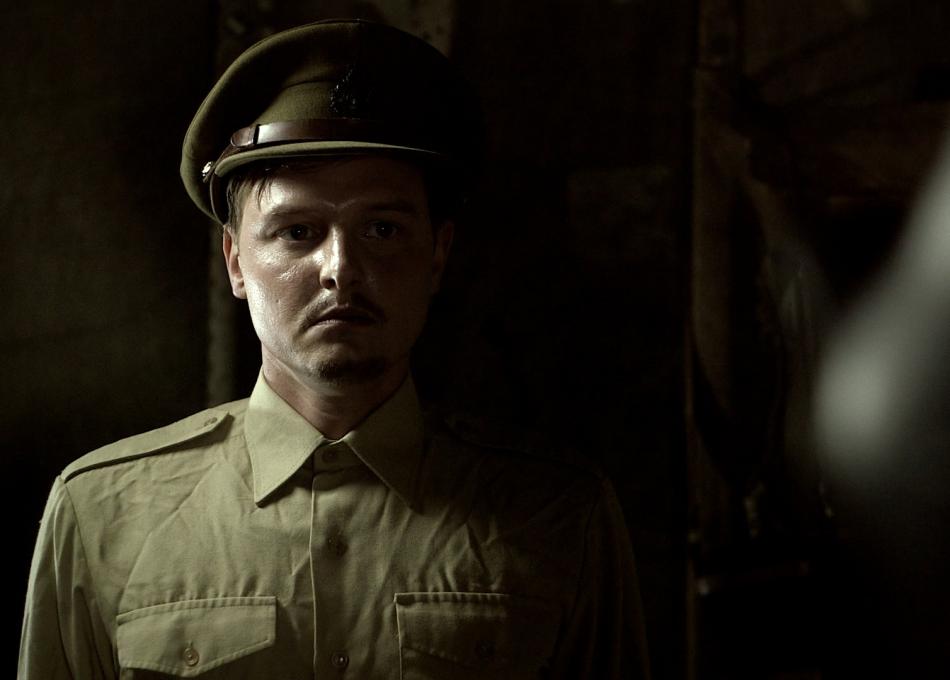 Serious-looking man in a khaki military uniform and cap, half his face in shadow.