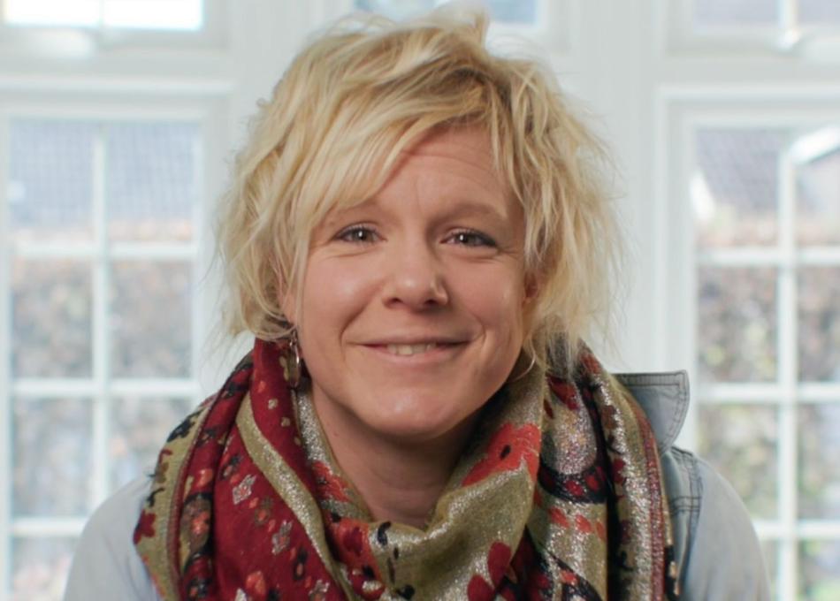 Photograph of a smiling woman with short blonde hair, wearing a colourful scarf, against blurred windows.