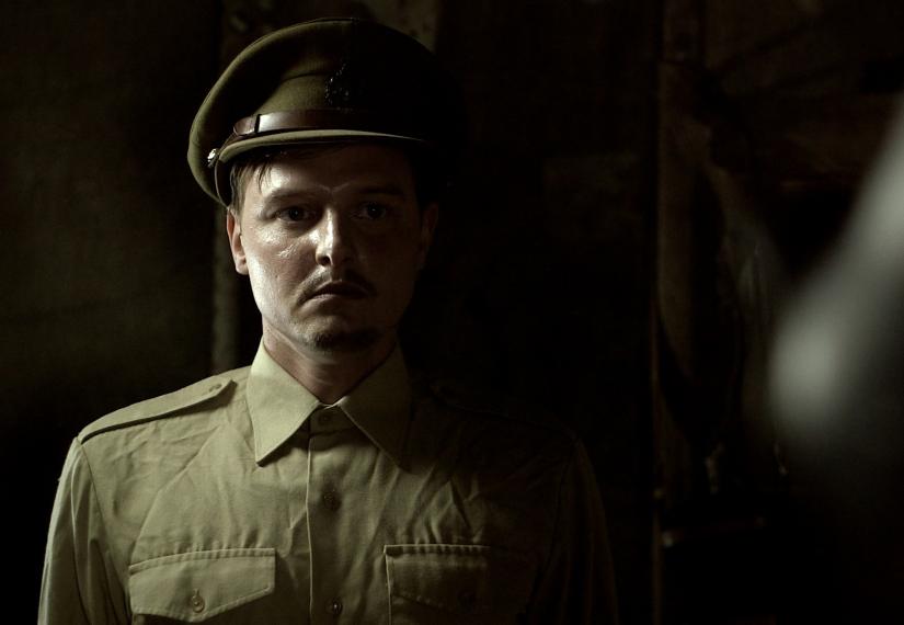 Serious-looking man in a khaki military uniform and cap, half his face in shadow.