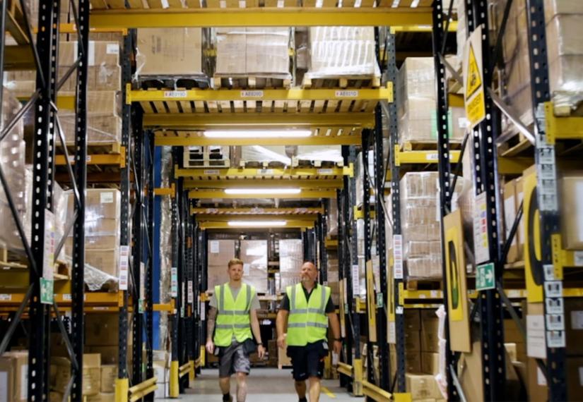 Two workers in high-vis vests walk down a busy warehouse aisle with tall shelves of goods.