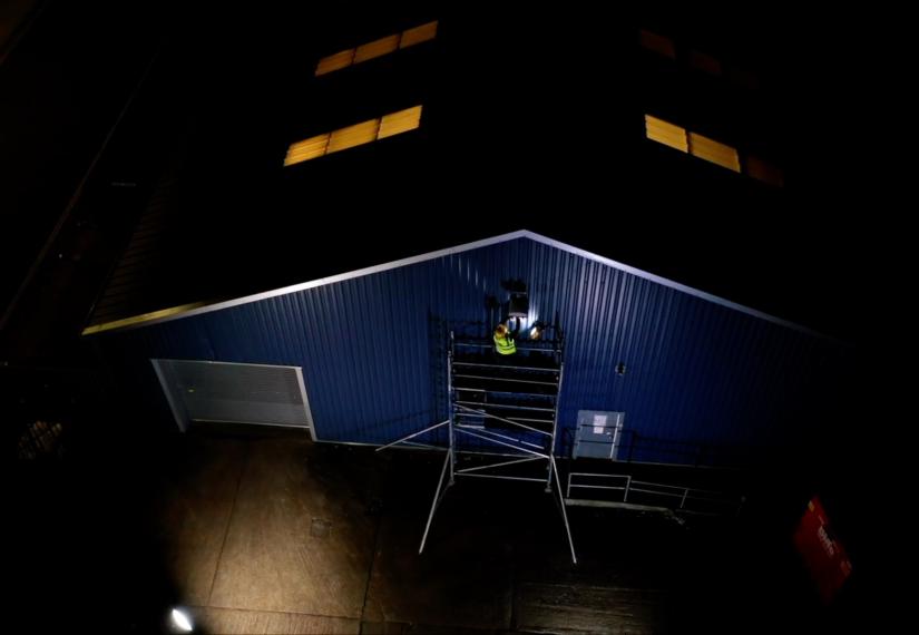 Dark blue building at night; worker on scaffolding near roof, illuminated windows above.