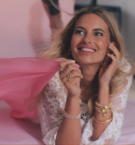 Smiling blonde woman in a lace top, lying on a pink studio background with gold jewellery.