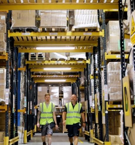 Two workers in high-vis vests walk down a busy warehouse aisle with tall shelves of goods.