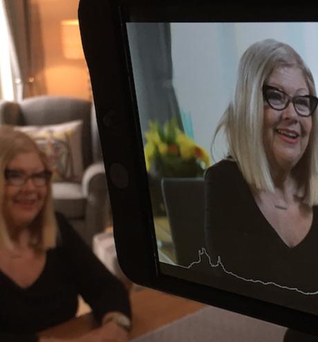 Lady being interviewed sat at table and seen on a camera monitor