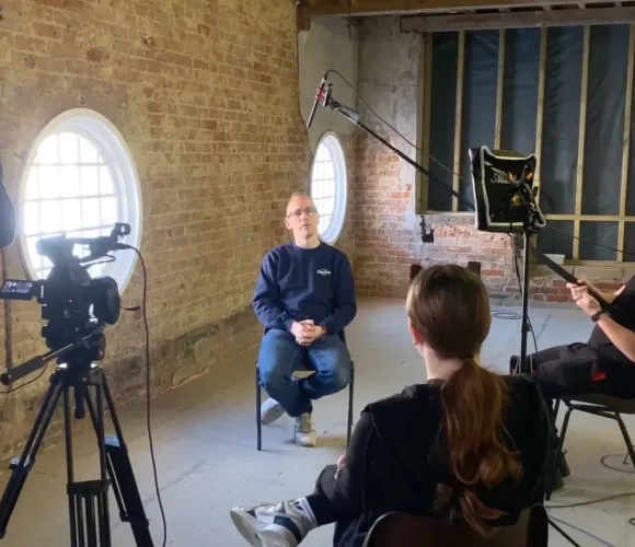 Film crew recording an interview in a brick-walled room with round windows.