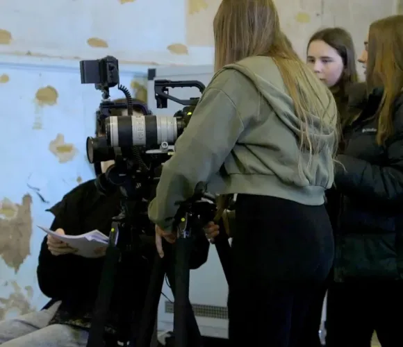 Three young women collaborate on a professional video camera setup; another person reads notes.