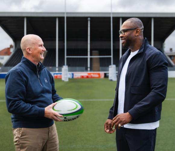 Two people talking and laughing whilst holding a rugby ball on a rugby pitch