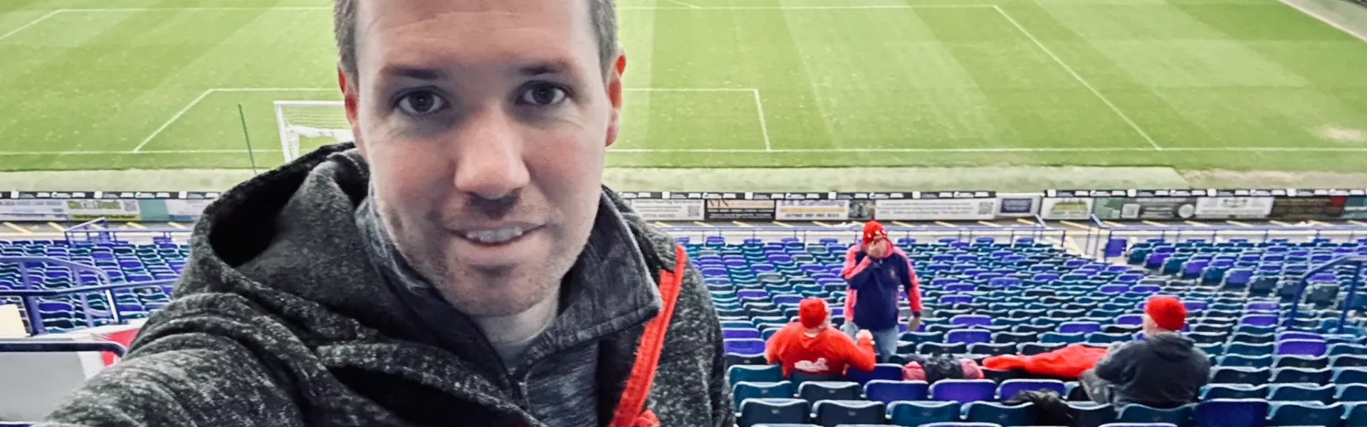 Man in a grey hoodie taking a selfie in a large, mostly empty football stadium.