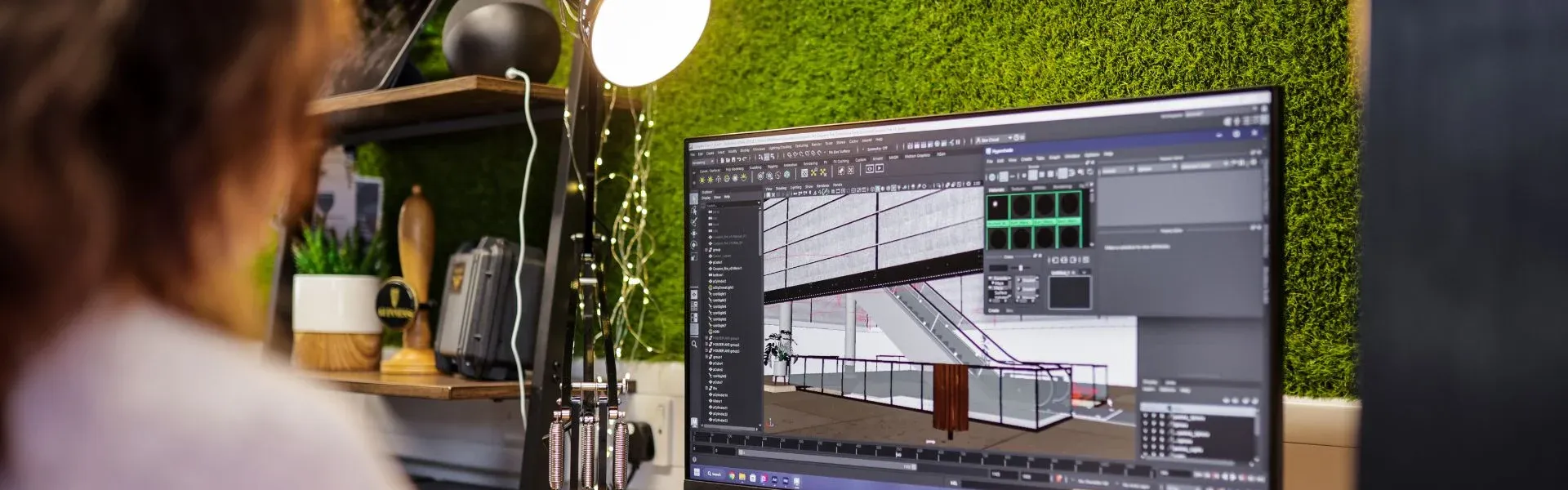 Designer focused on a monitor displaying 3D design software, green wall behind.