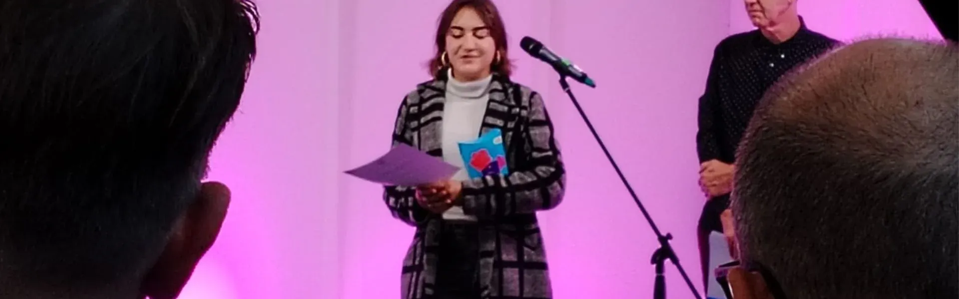 Woman smiling on stage, holding papers, with a man behind her. Pink lit background.