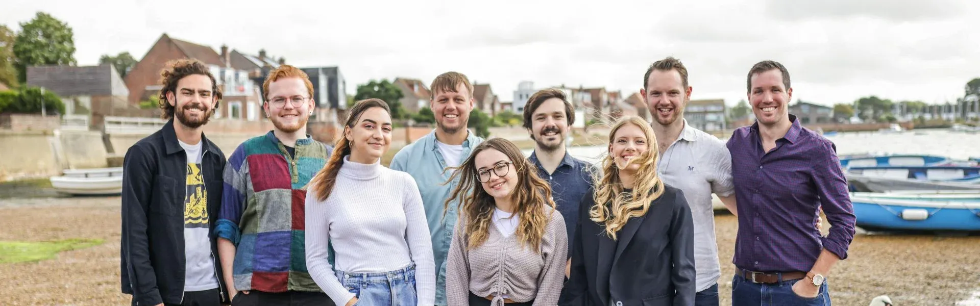 Photograph: Eight smiling people stand on a riverbank with boats and swans nearby.