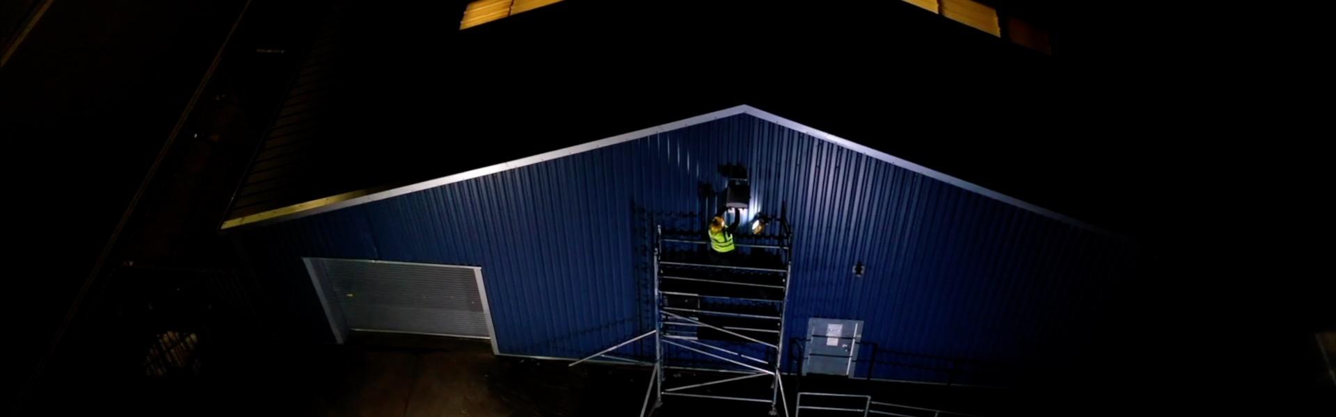Night-time photo of a worker on scaffolding against a dark blue building, windows glowing.
